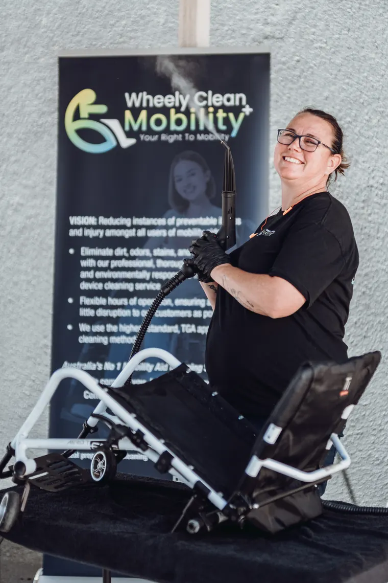 Kerry of Wheely Clean Mobility holding a steam cleaner beside a mobility walker, in front of the business banner.