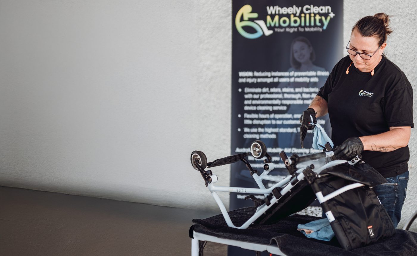 A Wheely Clean Mobility technician cleaning a folded mobility walker frame with a steam cleaner and microfibre cloth, in front of a Wheely Clean Mobility banner.